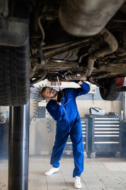 A female mechanic in blue coveralls inspects a car's undercarriage in an auto repair shop.